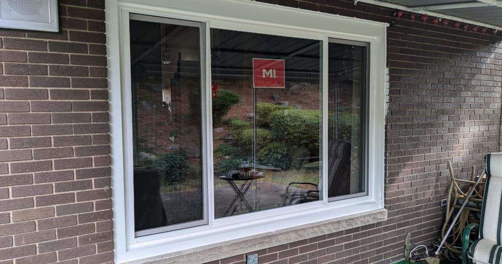 A white-framed sliding window on a brick building reflects a backyard with plants and furniture.