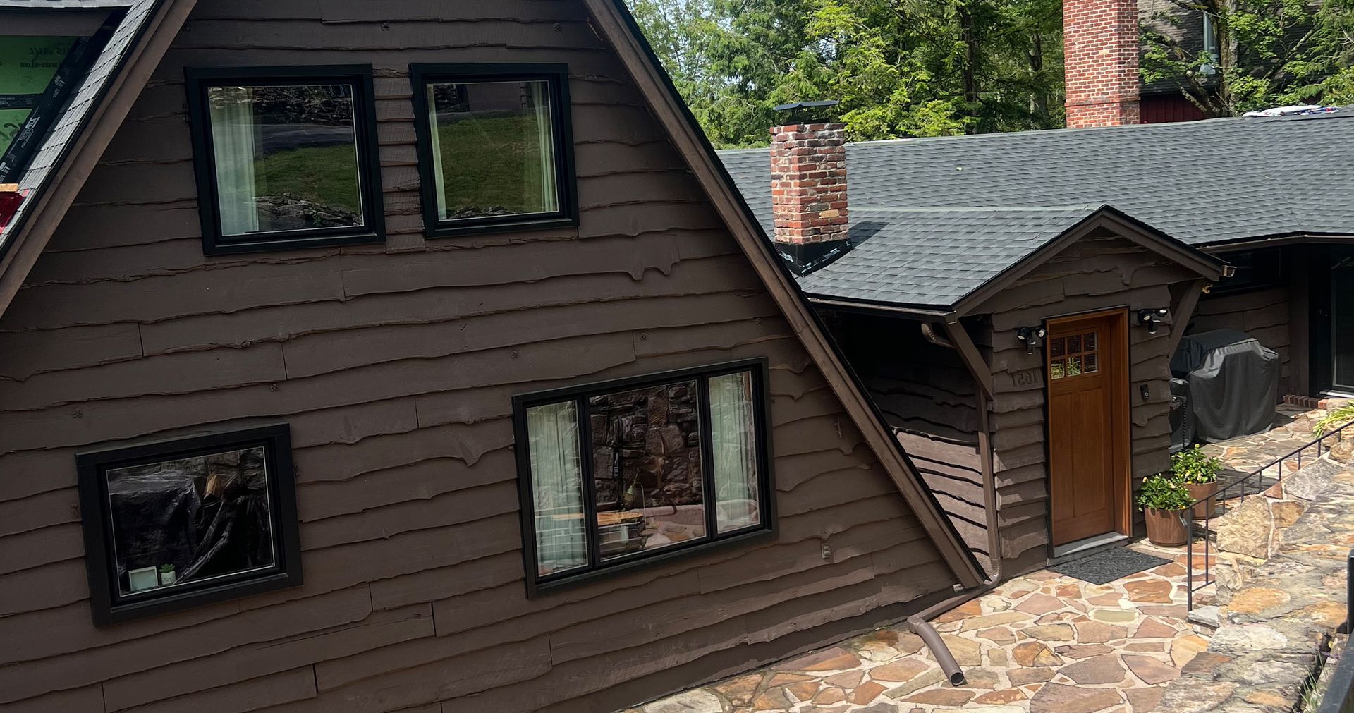 Brown A-frame house with black-framed windows, a brick chimney, and a stone pathway leading to the front door.