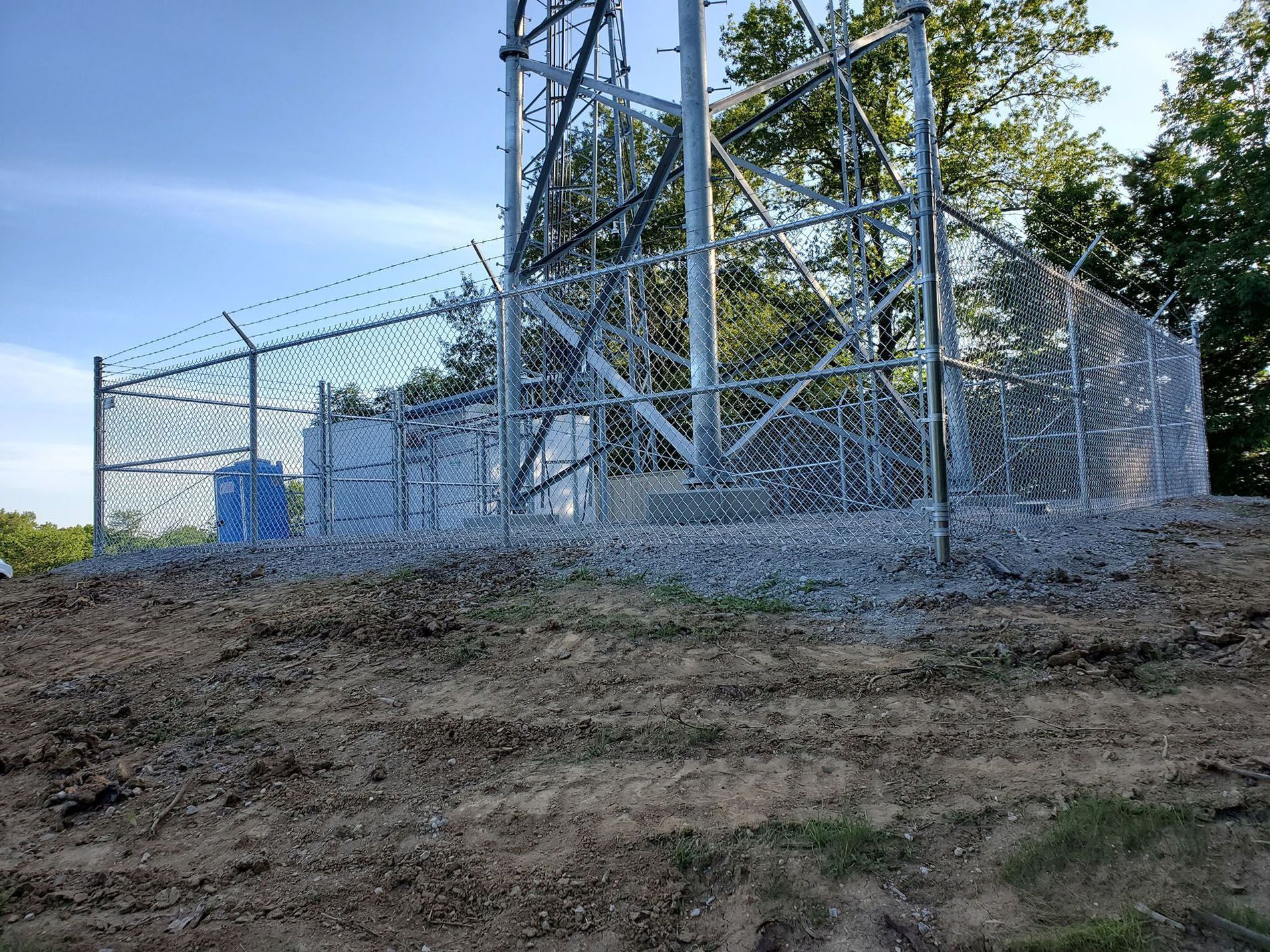 A chain link fence surrounds a power tower on top of a dirt hill.