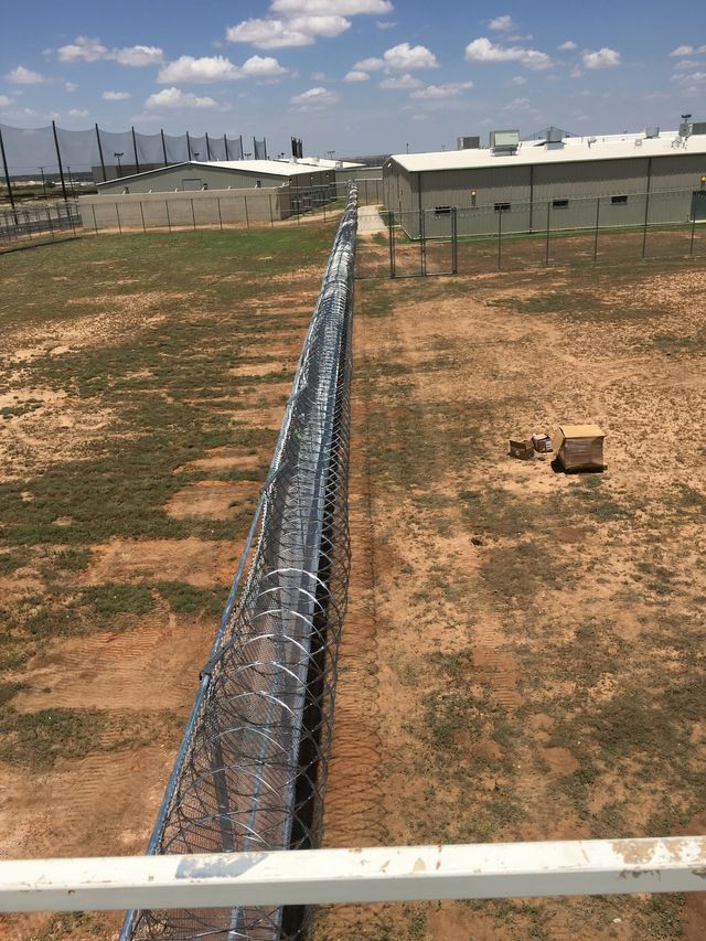 A barbed wire fence surrounds a dirt field with a building in the background.