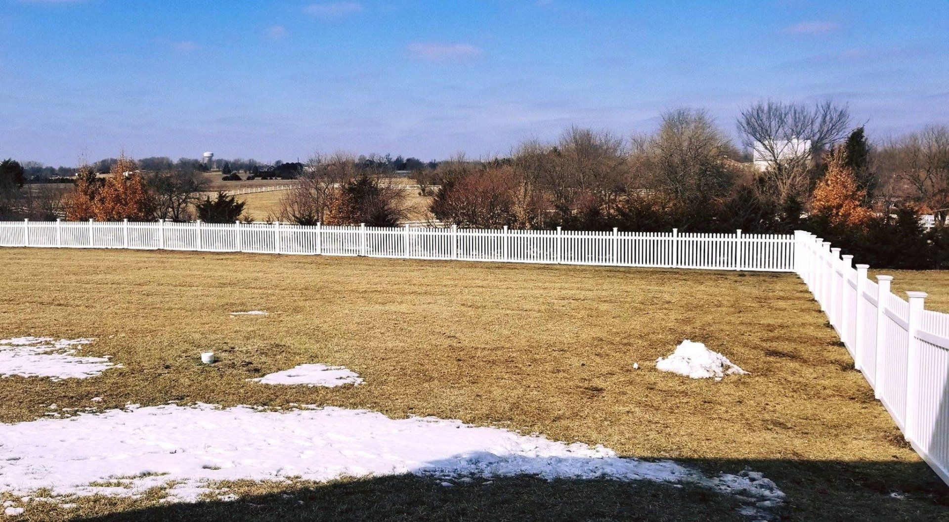 A white fence surrounds a grassy field with snow on the ground