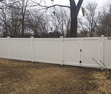 A white vinyl fence with a gate in a backyard with trees in the background.