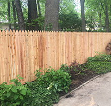 A wooden fence is surrounded by trees and bushes next to a sidewalk.