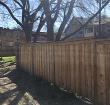 A wooden fence surrounds a yard with trees and a house in the background.