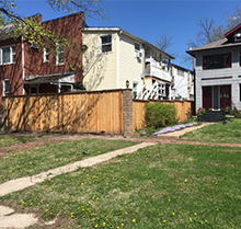 A wooden fence surrounds a lush green yard in front of a row of houses.