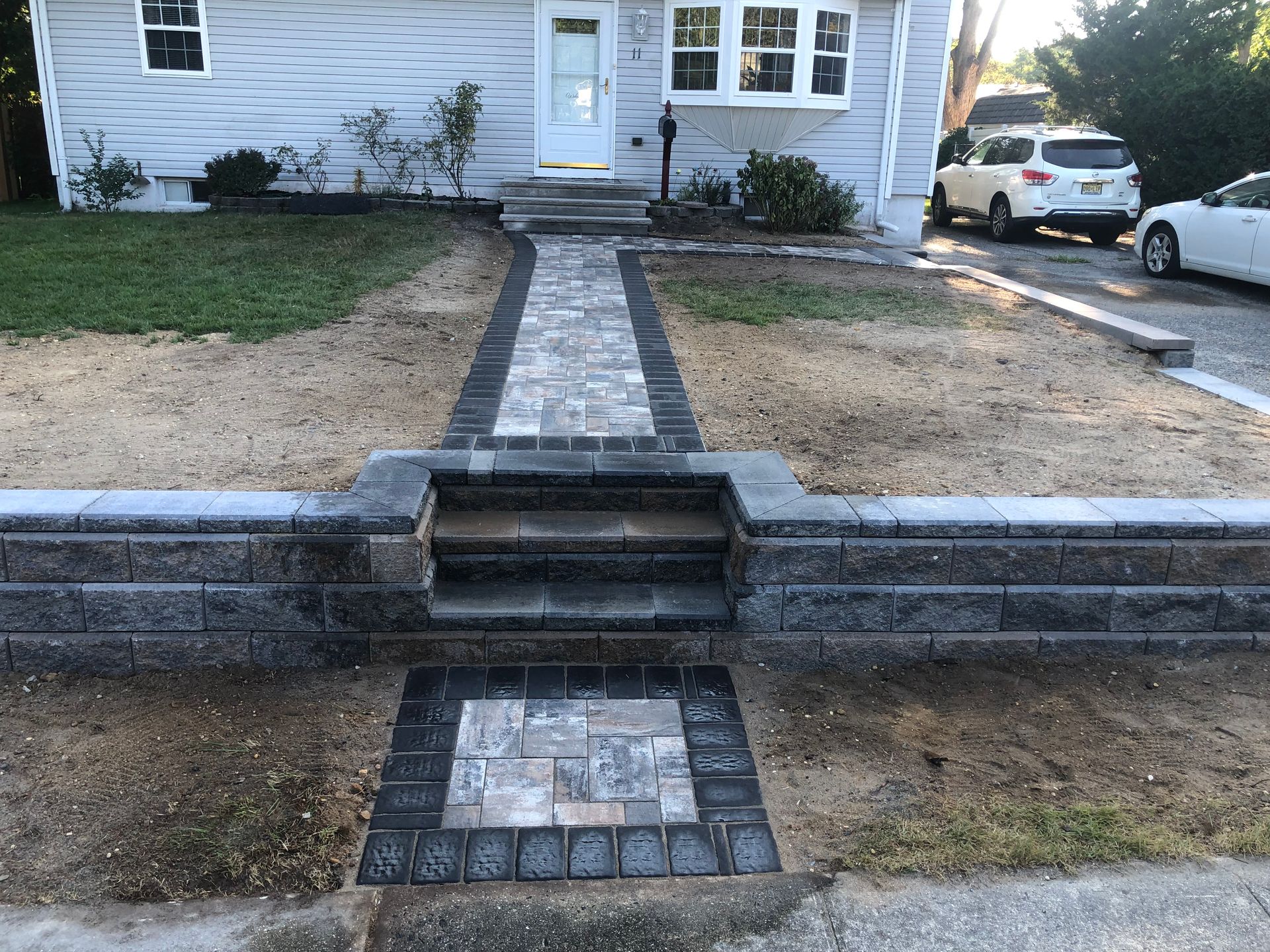 Brick walkway leading to a light gray house with front steps and retaining wall. Lawn and two parked cars are visible.