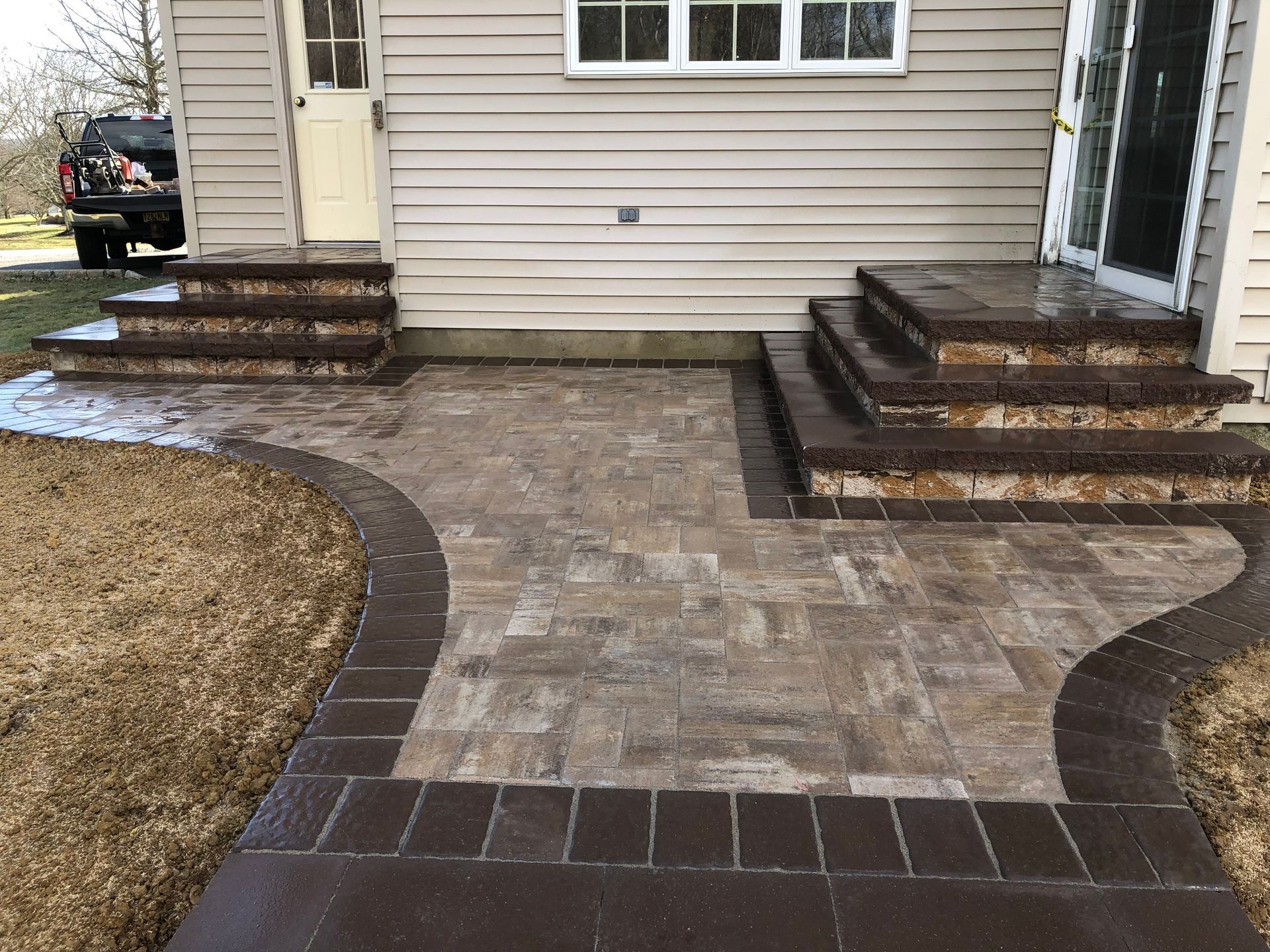 Brick patio with steps, bordered by darker bricks, leading to a house with siding.