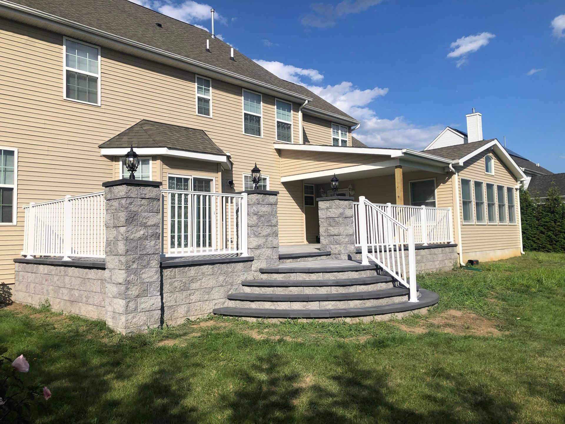 A multi-level backyard patio with steps, retaining walls, and white railing against a two-story house.