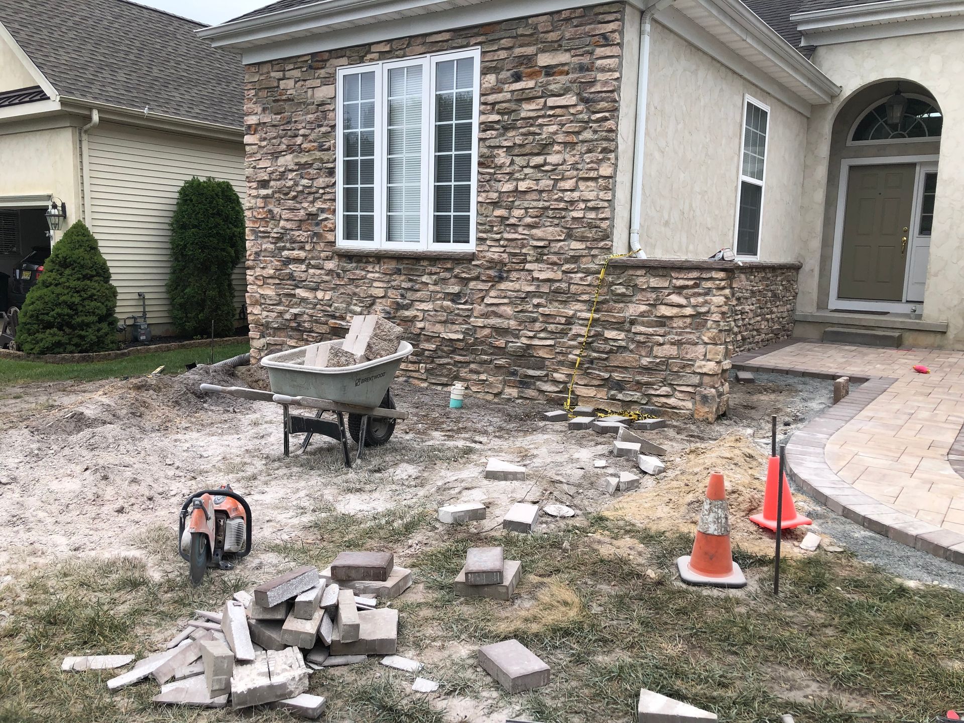 Construction site: stone veneer installation on a house exterior, bricks, wheelbarrow, tools, and grass.