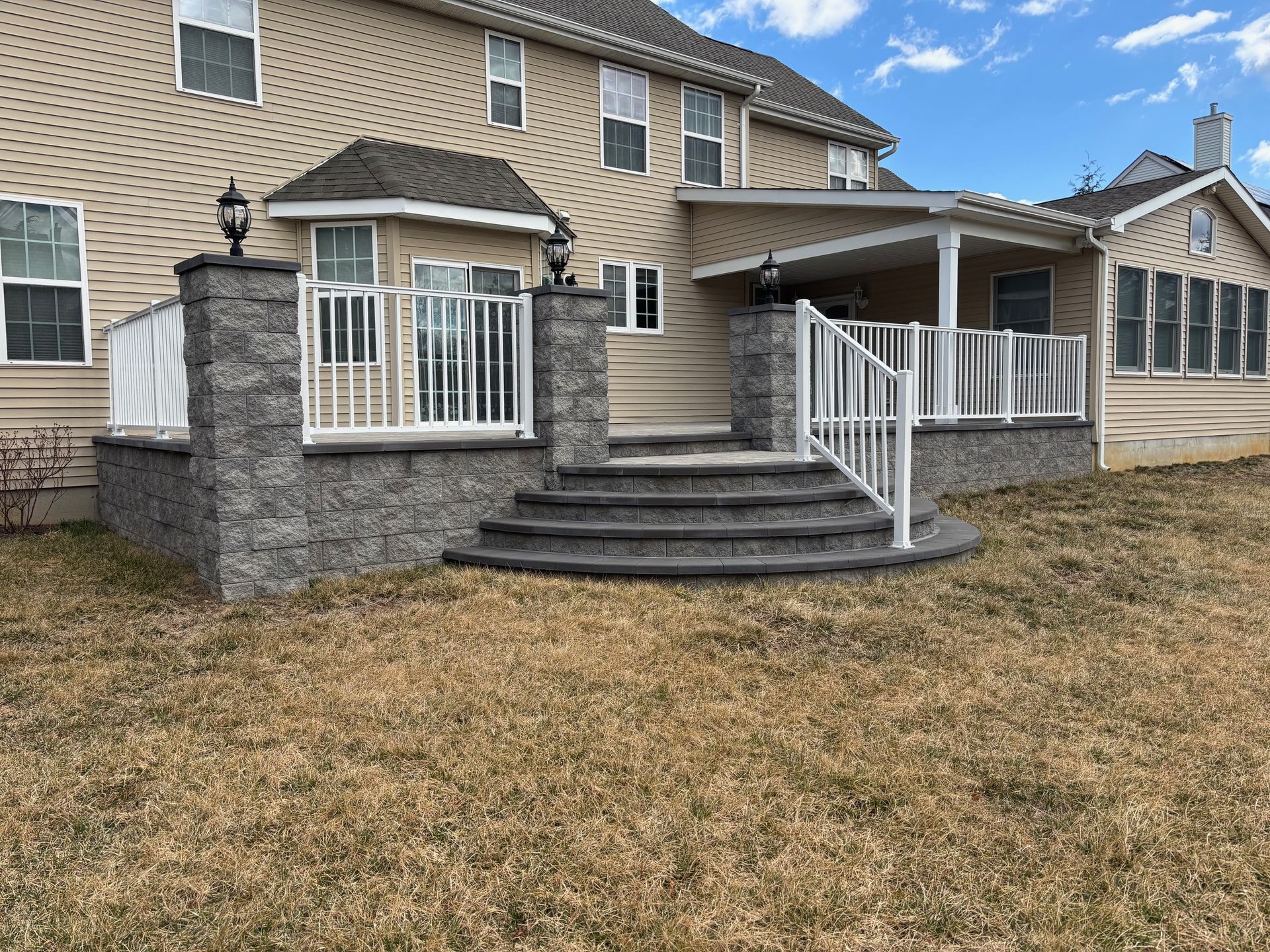 Backyard deck with stone stairs and railing; tan house in background, brown grass in foreground.