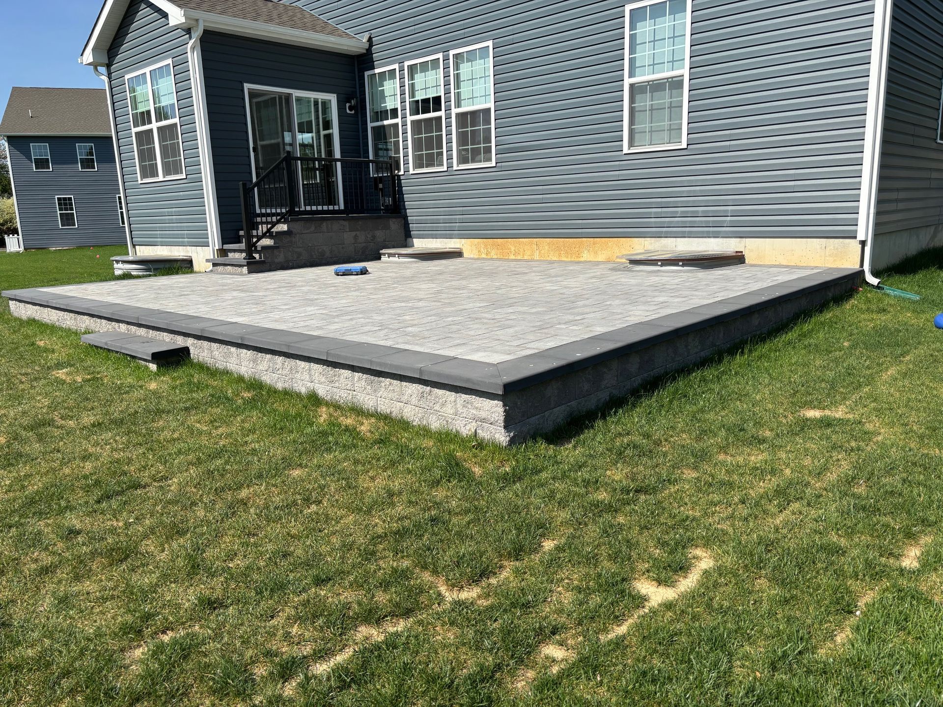 Backyard patio with grey pavers and a dark gray border next to a house with blue siding.