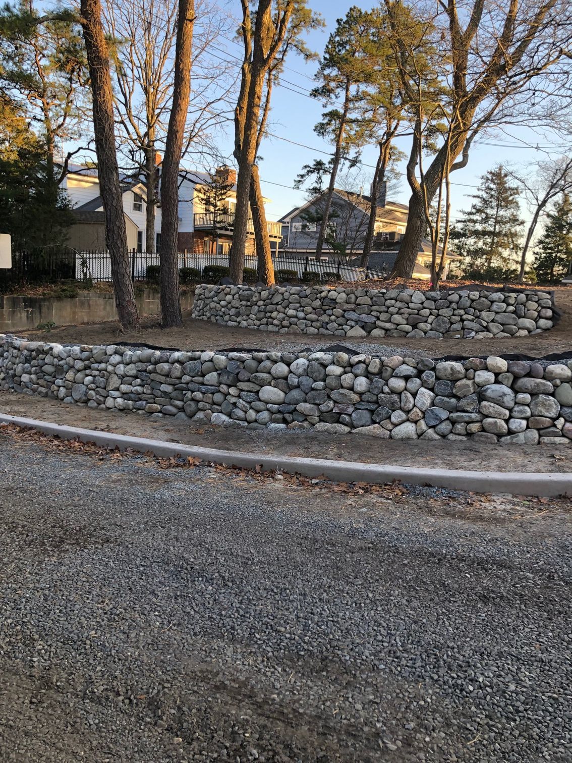 Stone retaining walls in front of trees and houses, gravel ground in the foreground.