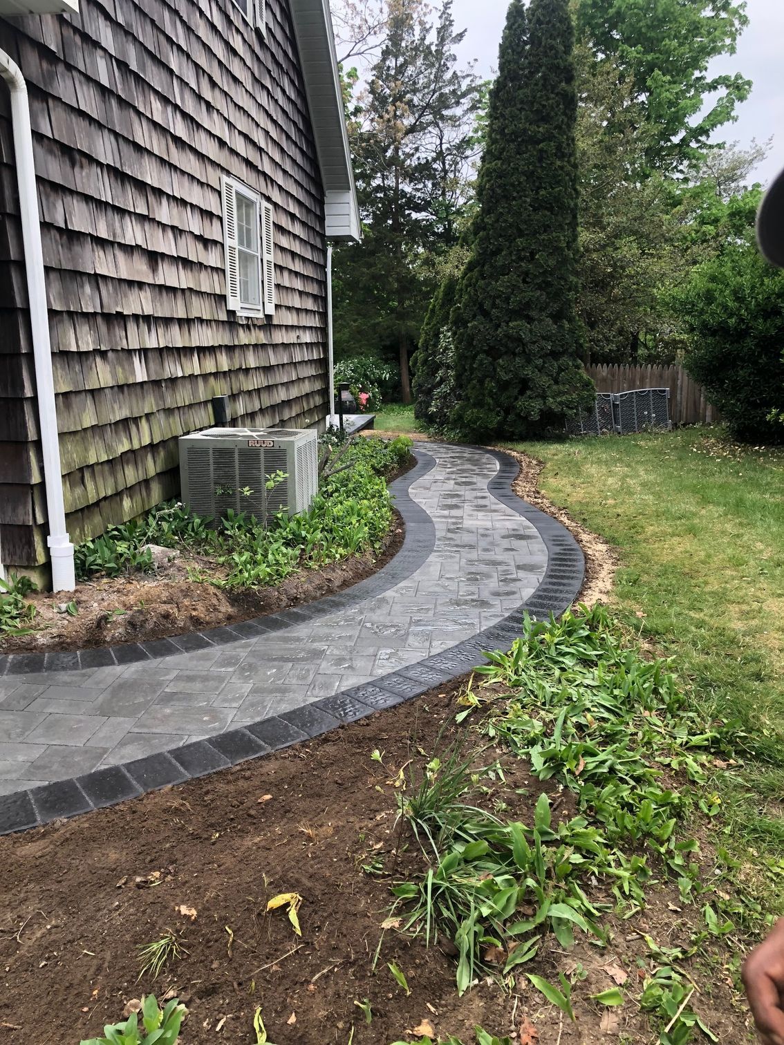 A winding gray and black brick pathway alongside a weathered wood-sided house and grassy yard.