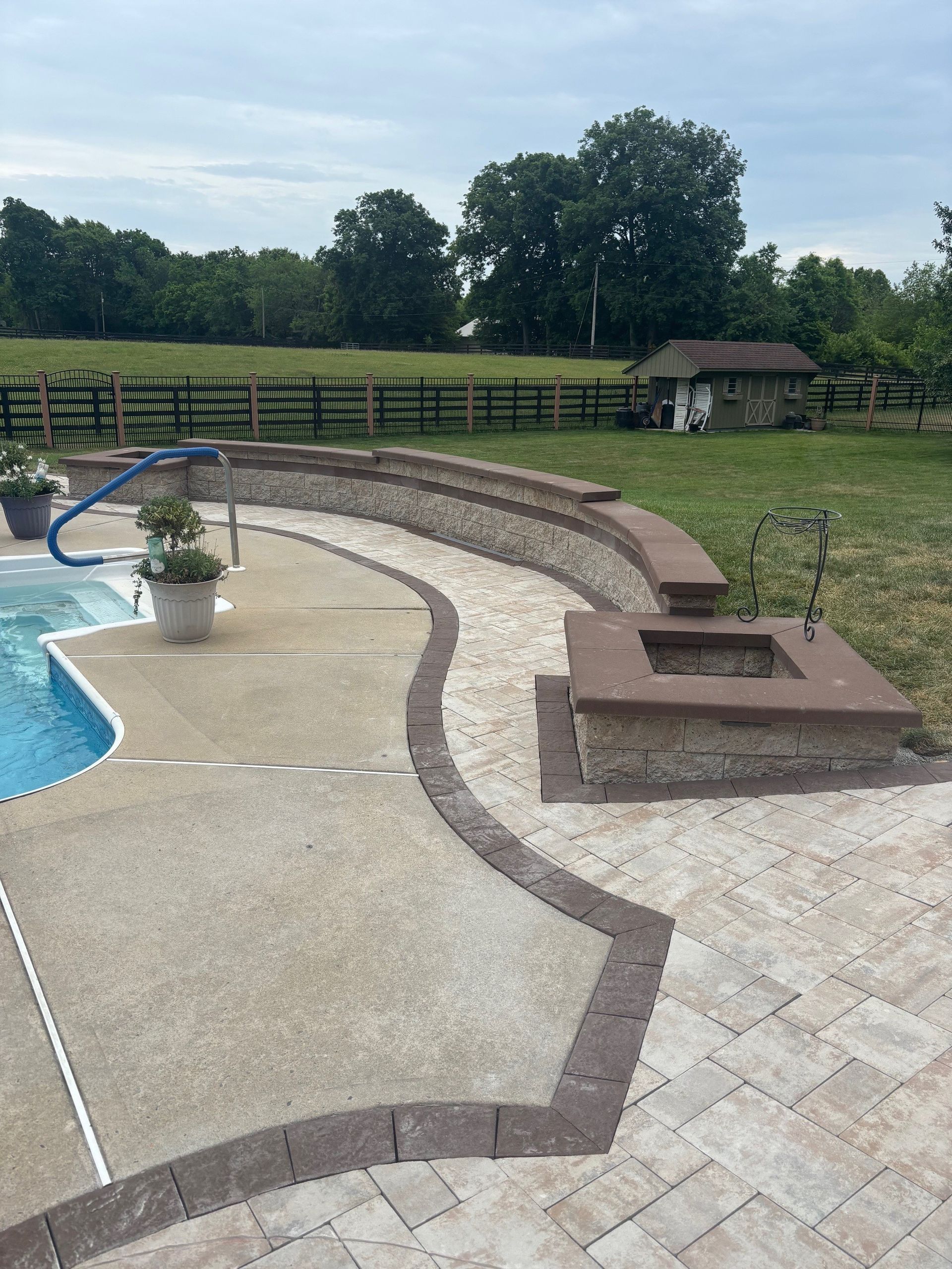 Backyard pool deck with curved stone seating, fire pit, and fenced grassy yard under cloudy sky