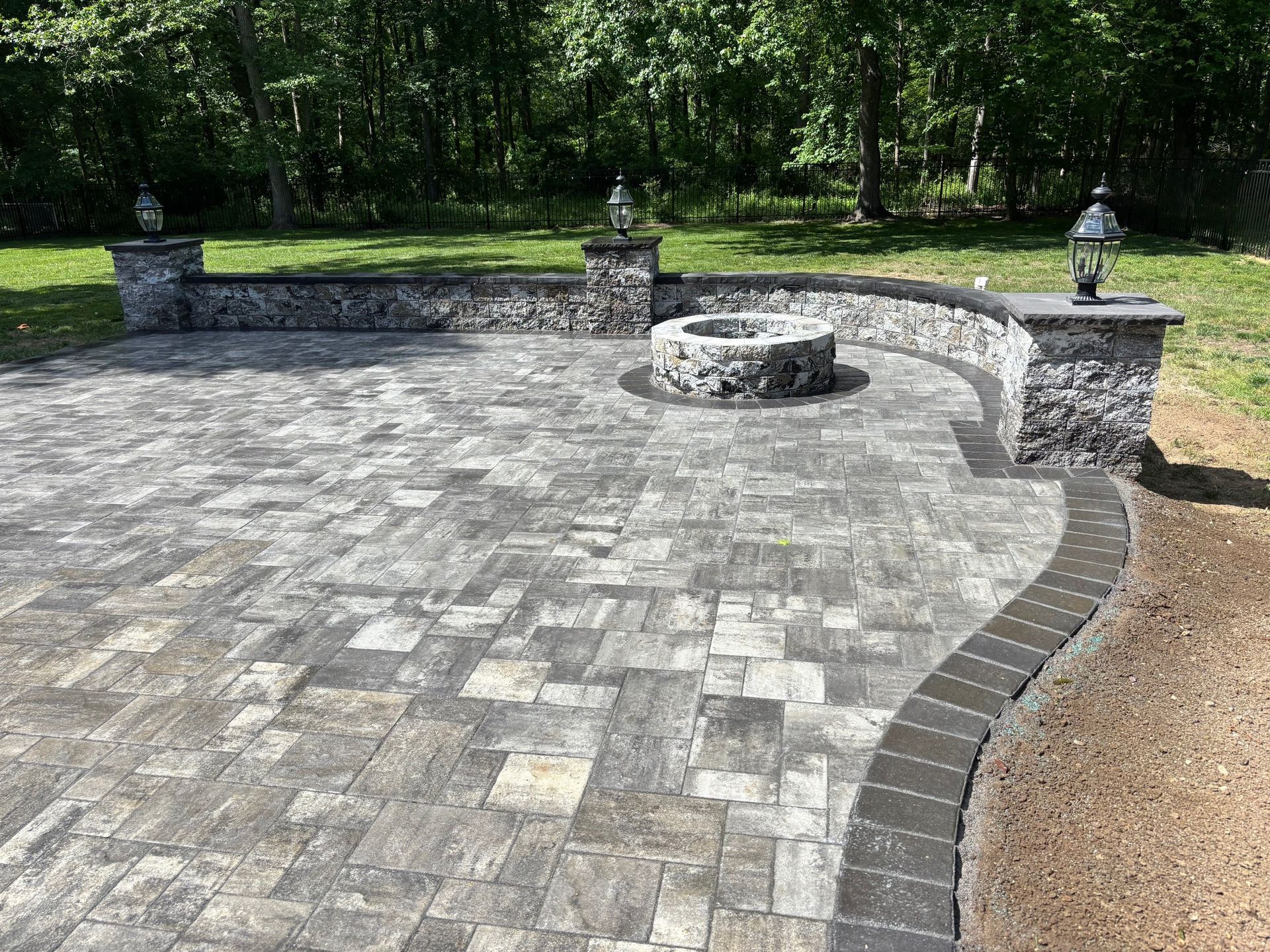 Stone patio with a curved low wall, central fire pit, and lantern posts beside a wooded area