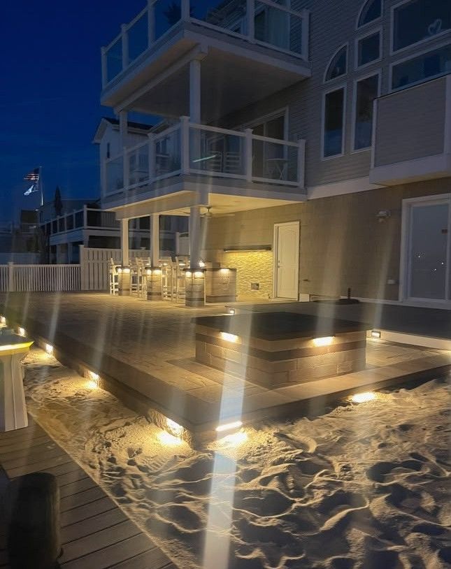 Lit deck lights beside a beach house at dusk, with sand in the foreground and lit balcony above.
