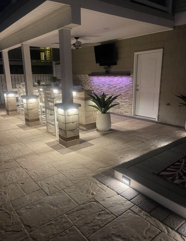 Lit patio with stone tiles, white columns, lantern lights, potted plants, and a wall-mounted TV beside a door.
