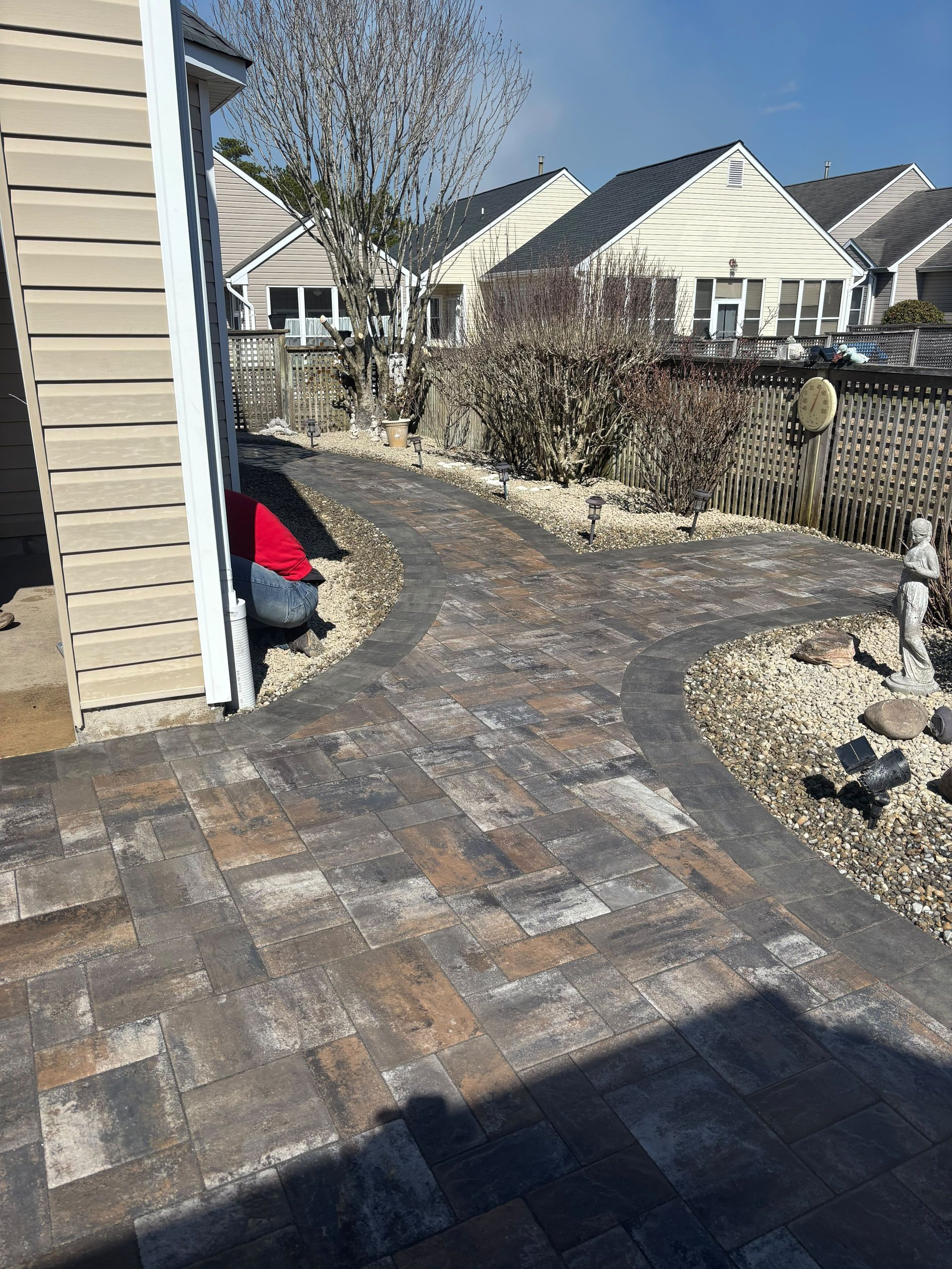 Curved brick patio path beside a house, with mulch beds, shrubs, and suburban homes in the background.