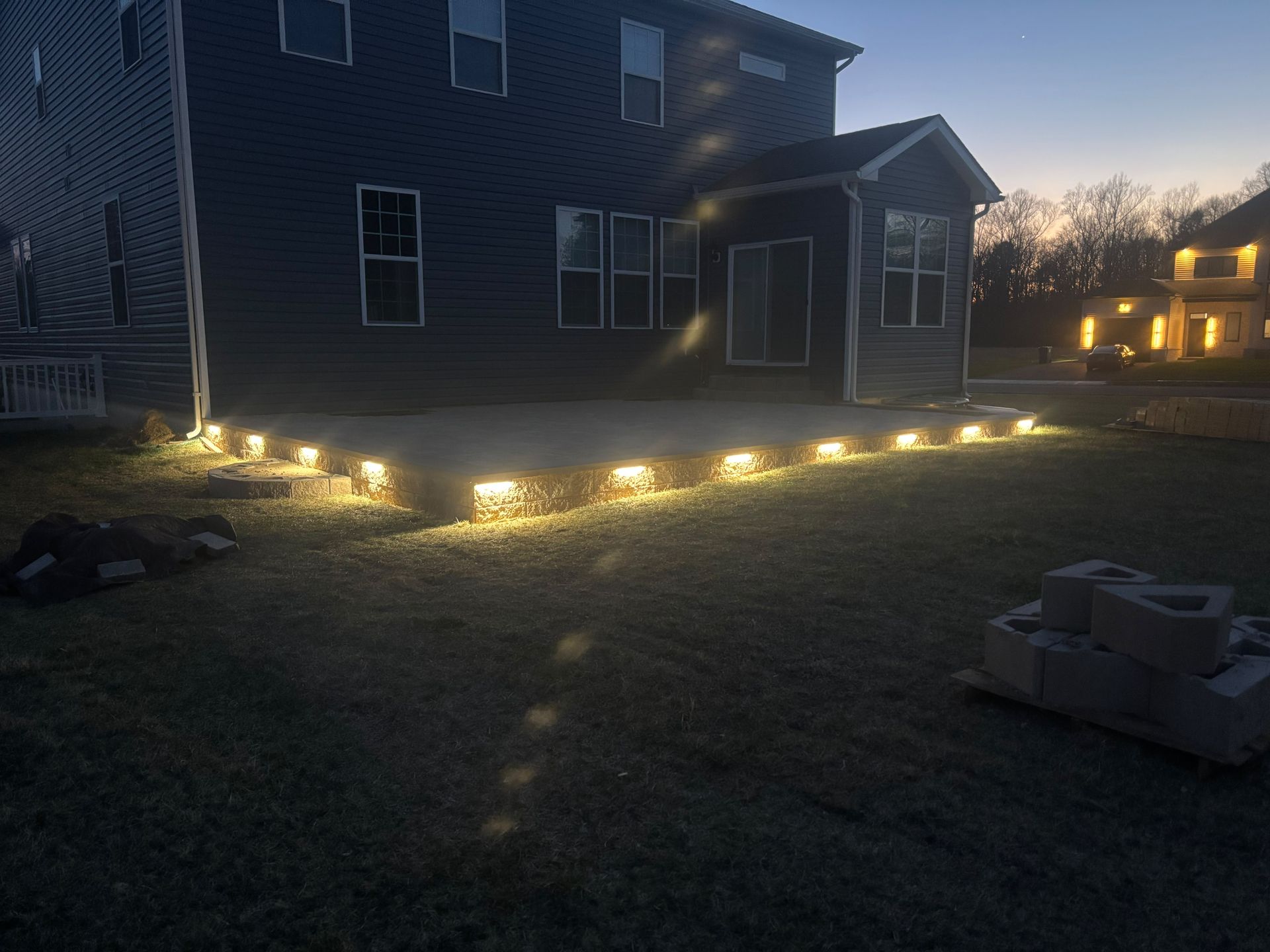 Backyard at dusk with house lit by warm ground lights along the patio and lawn.