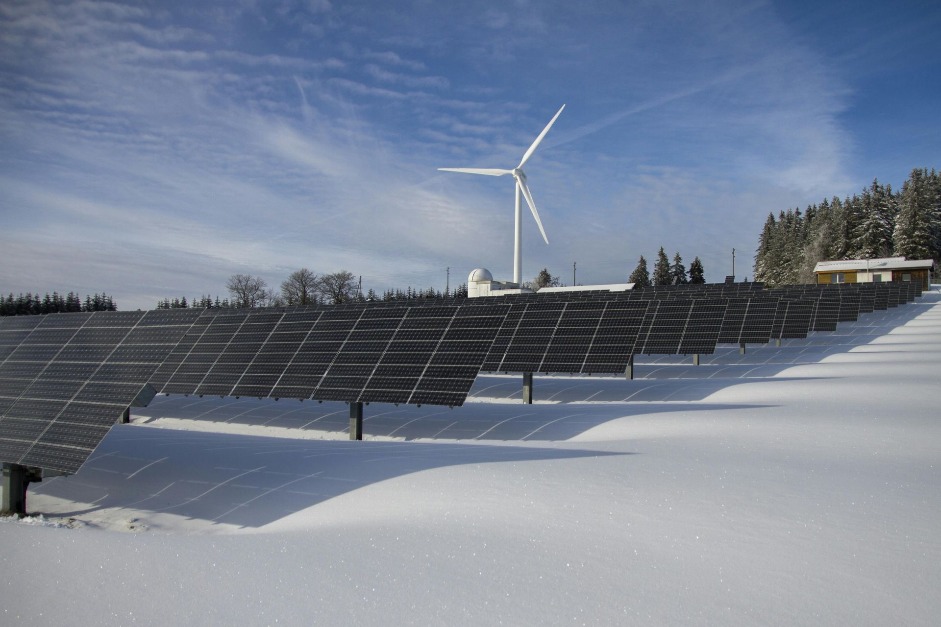 A row of solar panels in the snow with a wind turbine in the background.