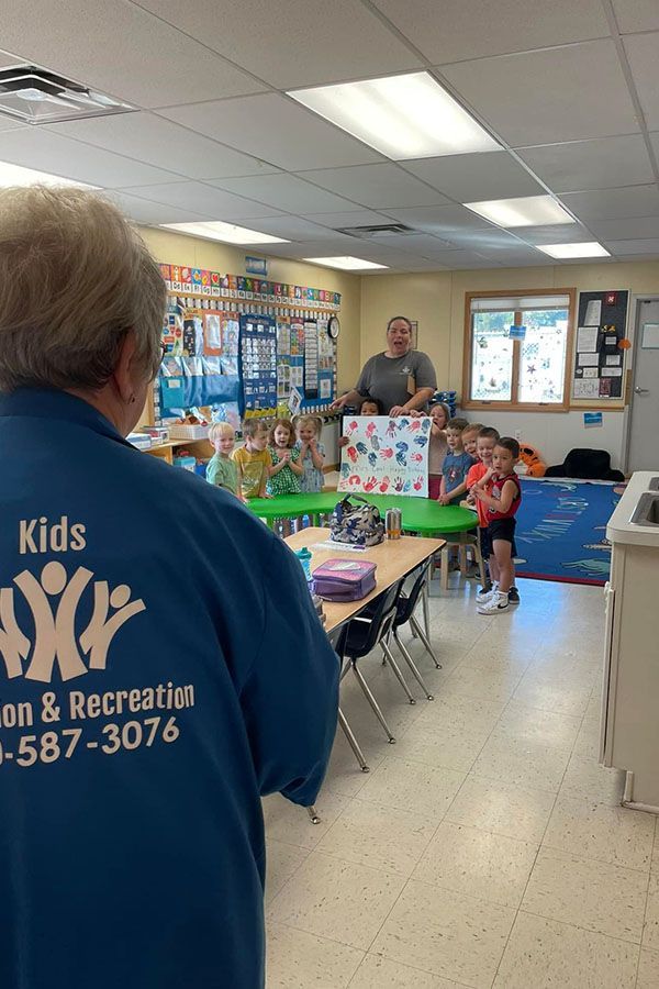 A man wearing a kids on & recreation shirt is standing in a classroom with children.