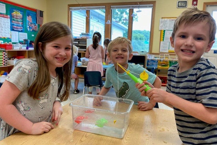 Three children are sitting at a table in a classroom playing with water toys.