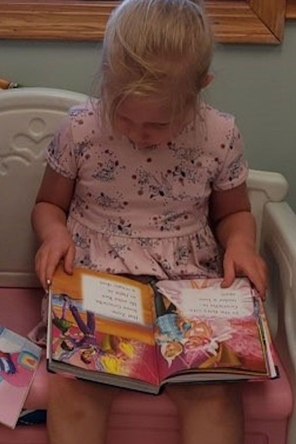 A little girl is sitting on a pink chair reading a book.
