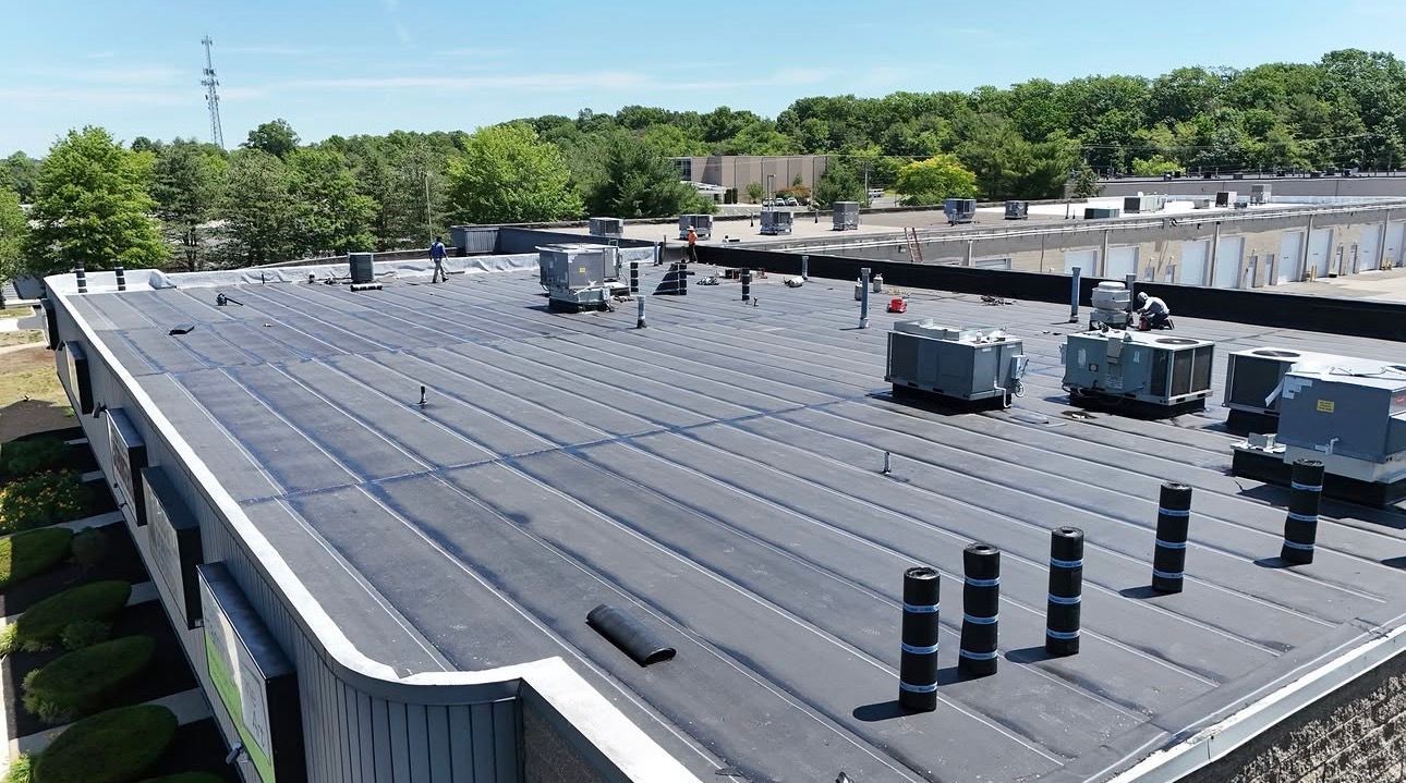 Flat commercial rooftop with HVAC units, vents, and a dark membrane roof under a blue sky.