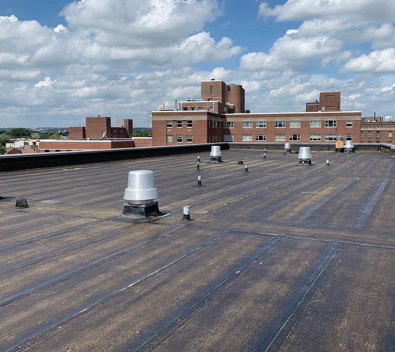Flat rooftop with vents and a brick building under a partly cloudy sky.