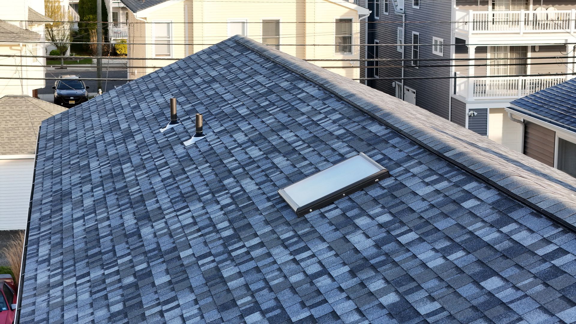 Gray shingled rooftop with vents and a skylight, surrounded by neighboring buildings.
