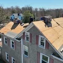 Roofers replacing shingles on a multi-story house with exposed plywood roof sections.