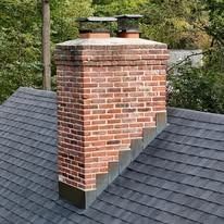 Brick chimney on a gray shingled roof with a metal cap, surrounded by trees.