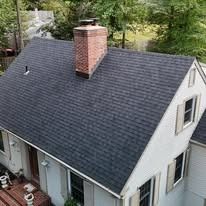 Aerial view of a gray shingled house roof with a brick chimney and dormer windows surrounded by trees