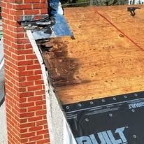 Partially shingled roof with plywood sheathing and black underlayment beside a brick chimney