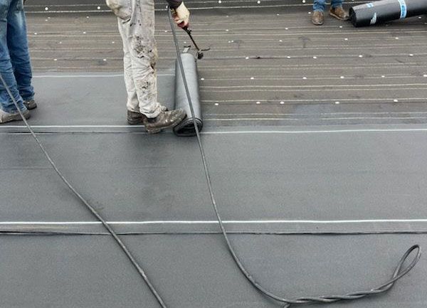 Workers installing black roofing material on a flat roof with loose cables and tools nearby