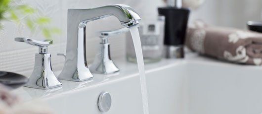 A chrome bathroom faucet with two handles pouring water into a white sink, with toiletries in the background.