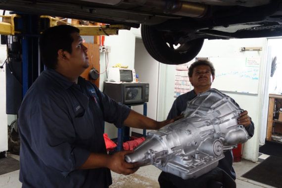 Two men are holding a large piece of metal under a car