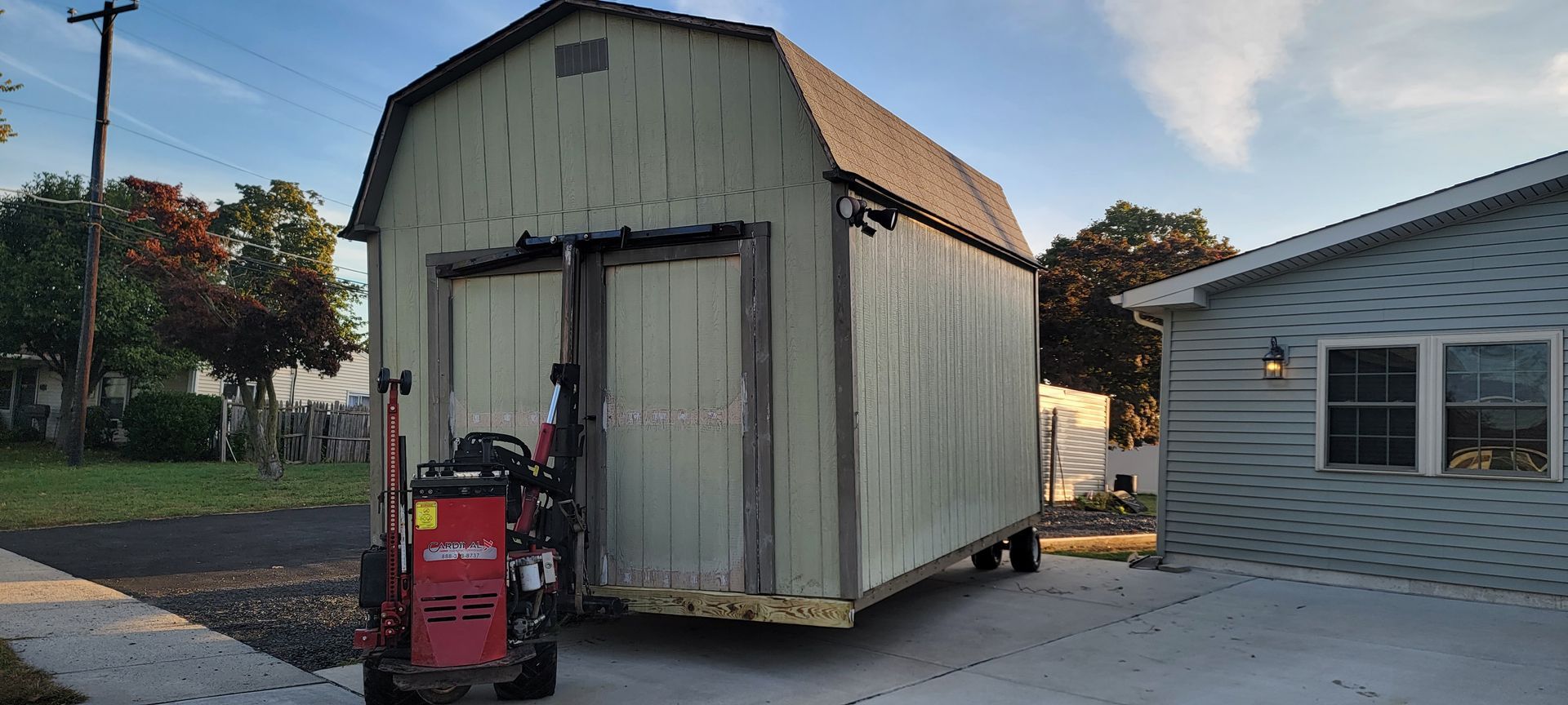 A shed is being lifted by a forklift in front of a house