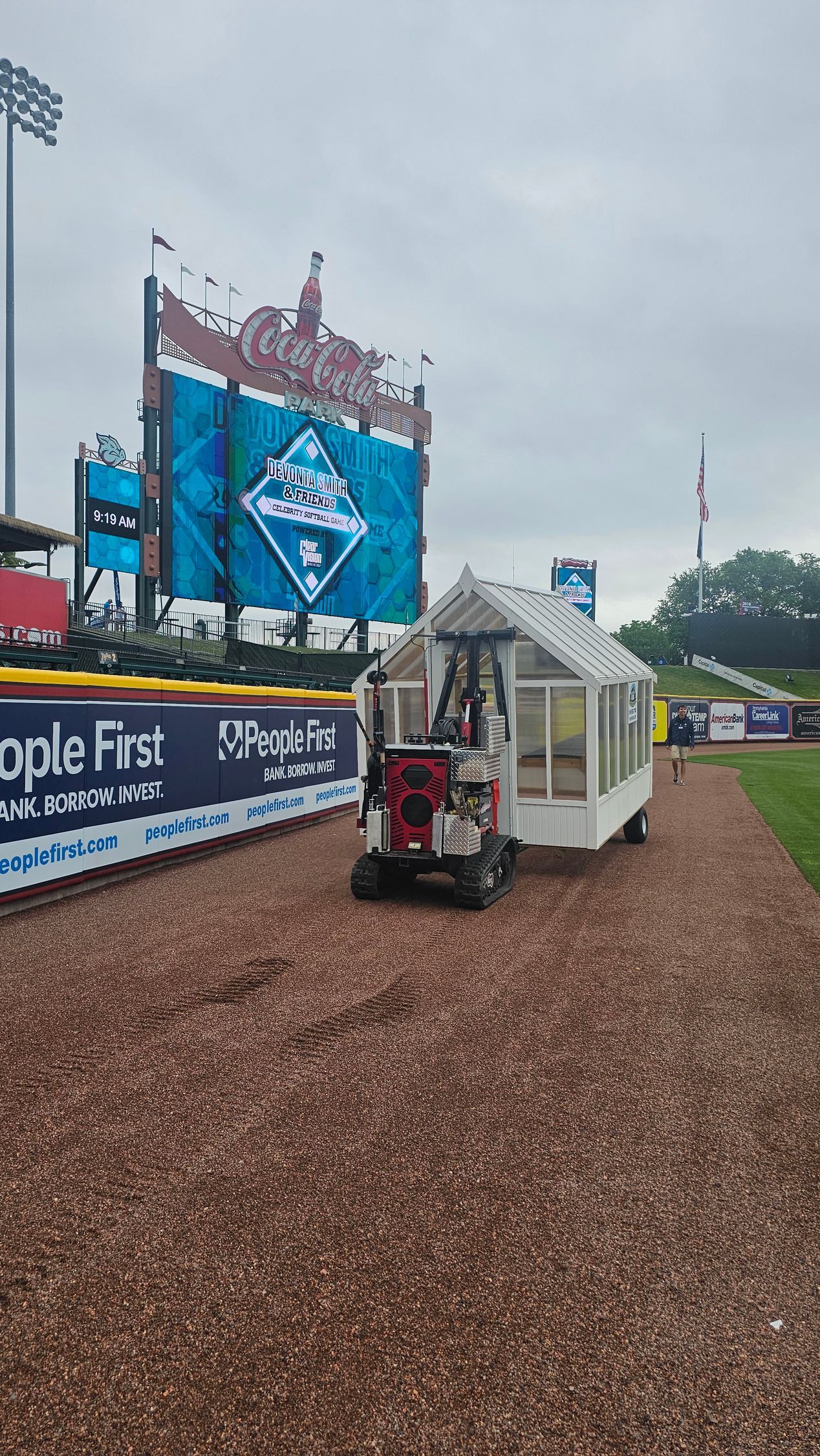 A tractor is pulling a greenhouse on a trailer on a baseball field.