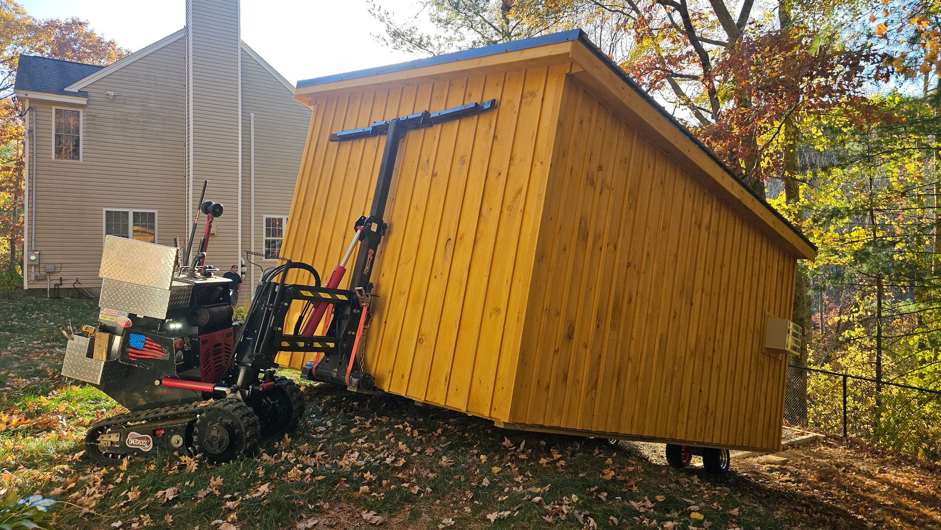 A yellow shed is being lifted by a forklift in front of a house.