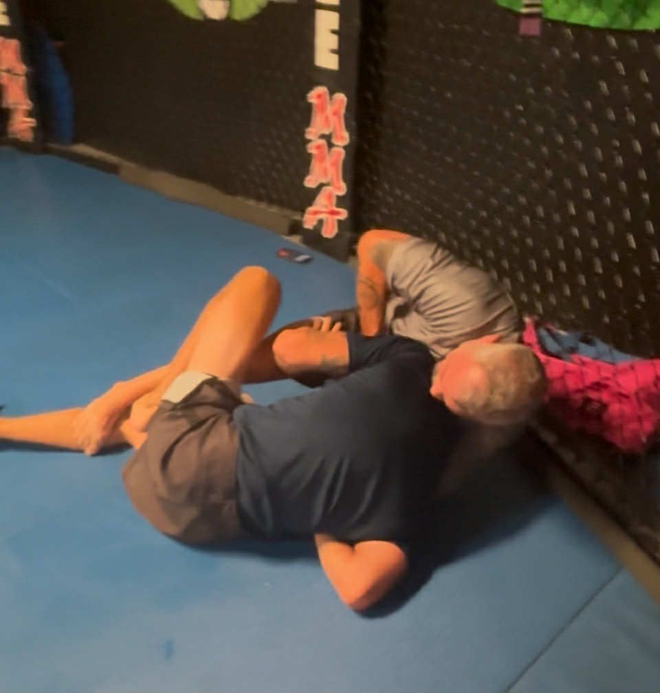 Two men grappling on a blue mat inside a gym. One is on his back, legs entangled with the other, who is partially visible.