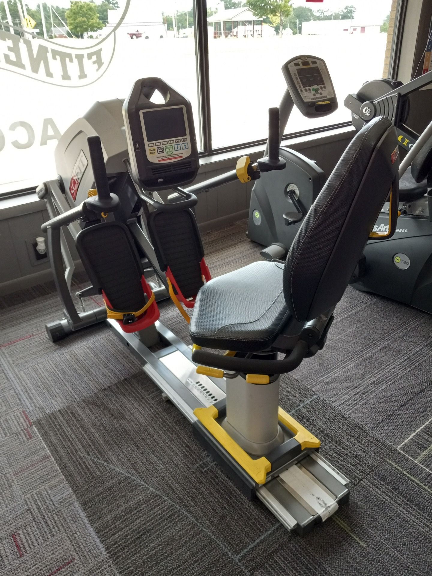 Recumbent exercise bike in a gym. Gray and yellow frame, black seat, with a digital display.