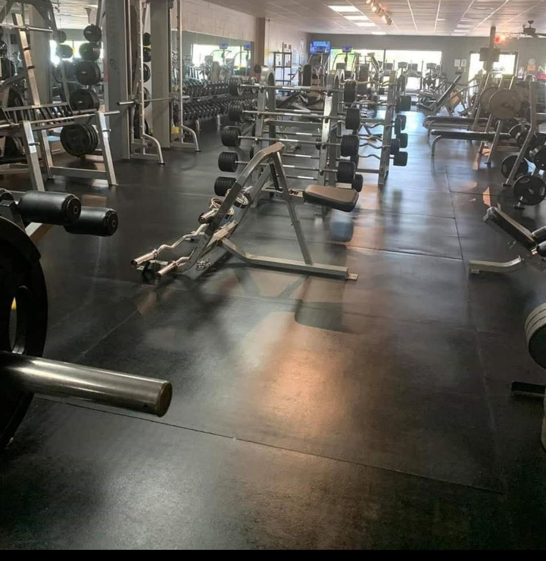 Gym interior with weight racks, machines, and free weights on a black floor. Rows of equipment fill the space.