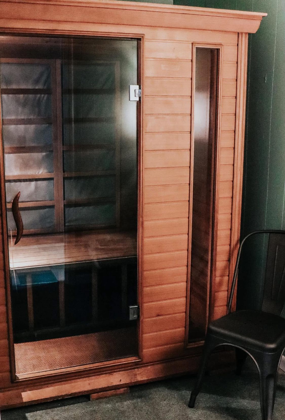 A wooden sauna with a glass door, a black chair sits beside it against a green wall.