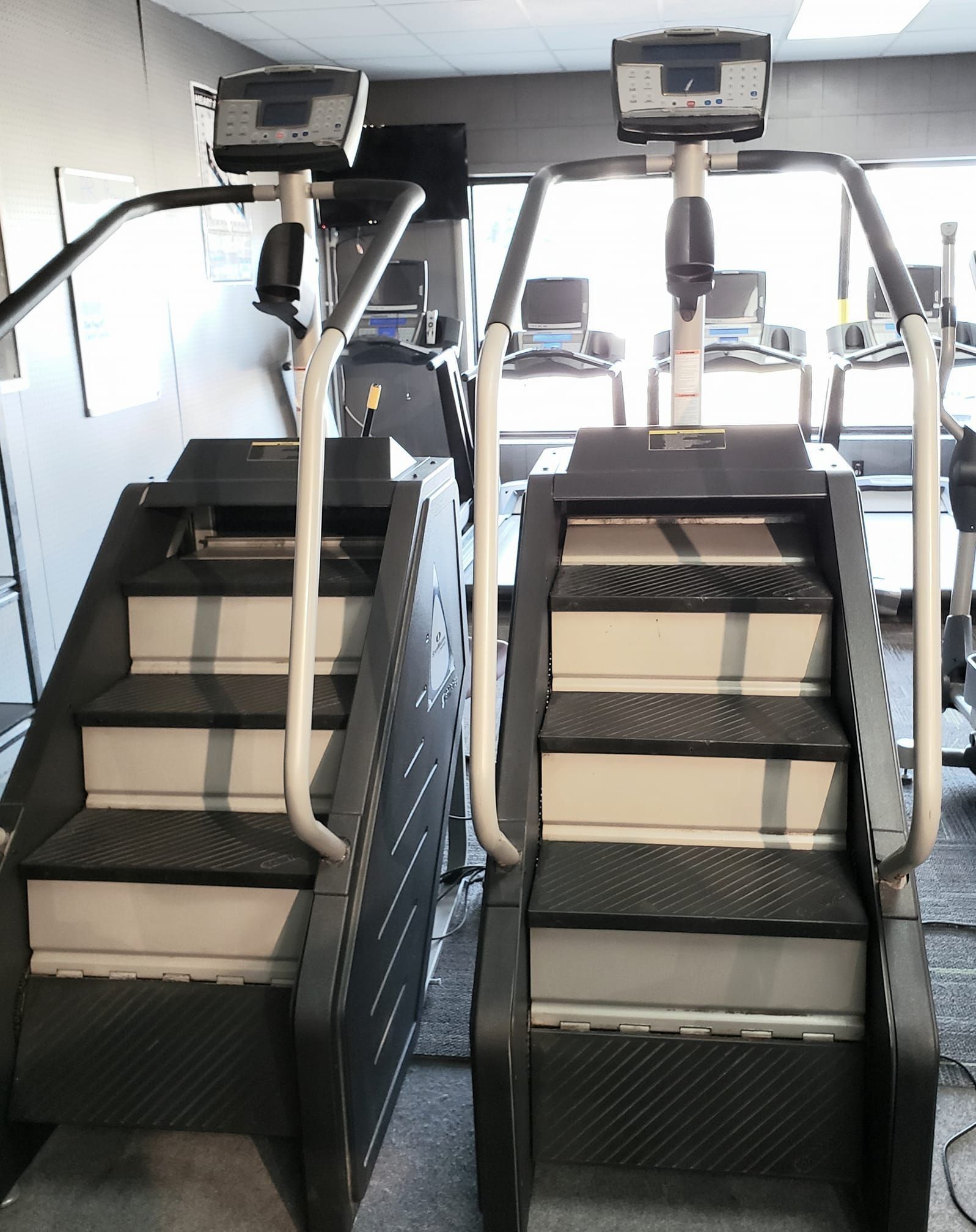 Two stair stepper machines in a gym. They have black steps, white railings, and digital displays at the top.