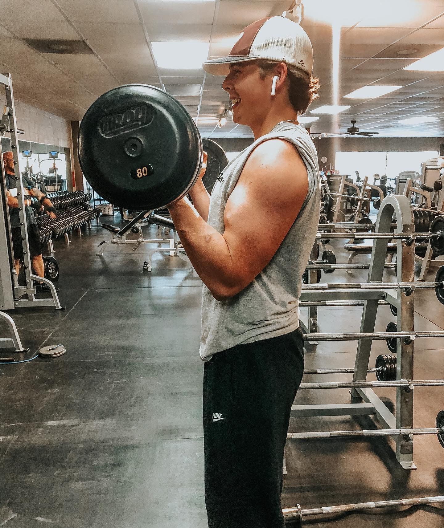 Man in a gym does a bicep curl with a barbell. He wears a hat, gray tank top, and smiles. The gym has various weight machines.