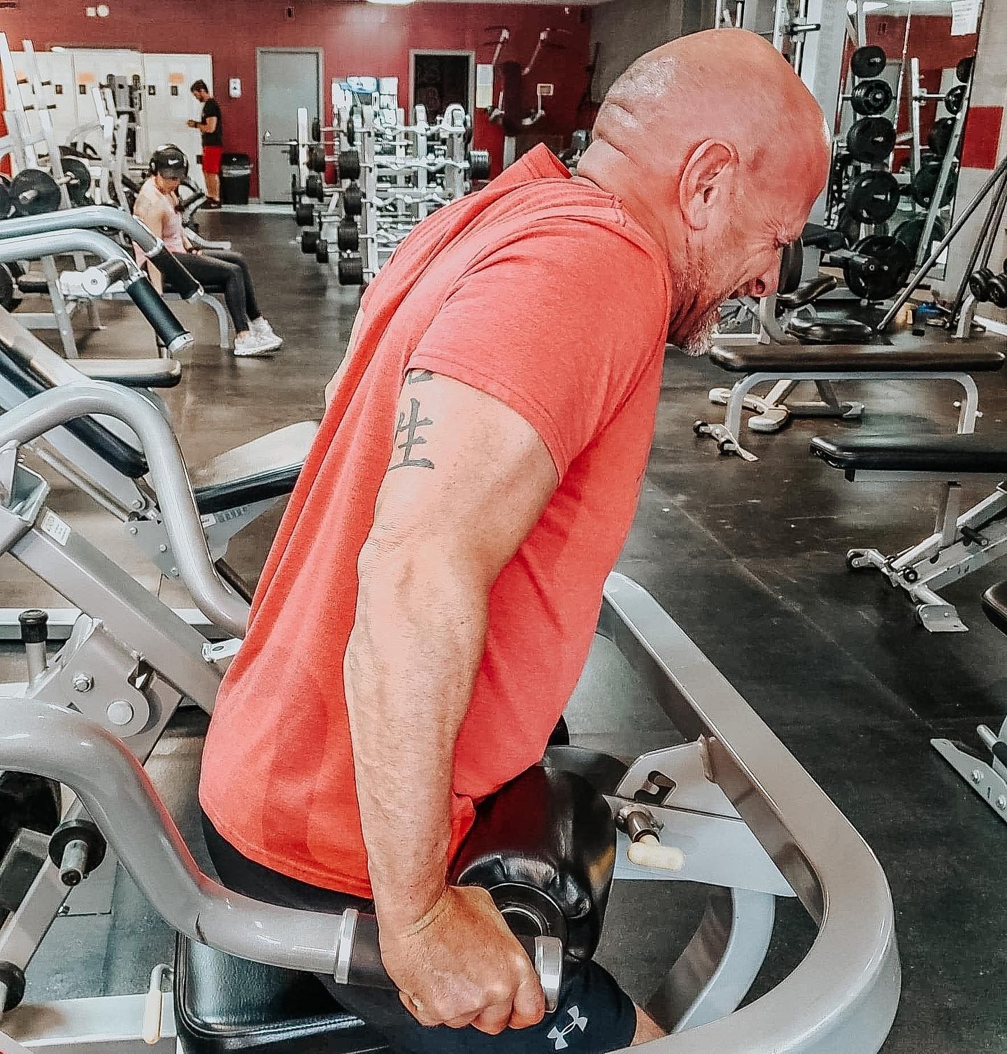 A bald man in a red shirt is bent over, lifting a weight machine handle at a gym. He appears to be straining during the exercise.