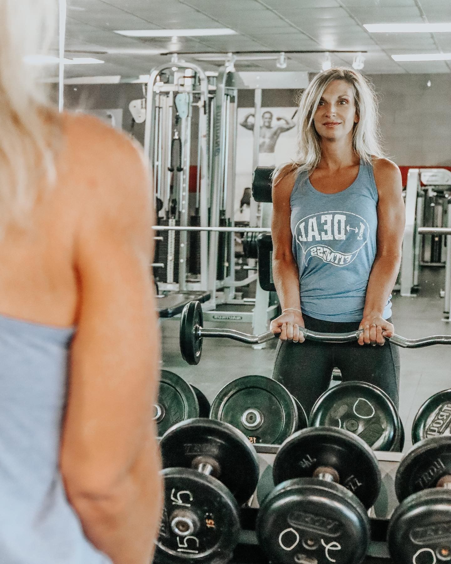 Woman in a gym lifting weights, reflected in a mirror. She wears a blue tank top and black leggings, with a serious expression.
