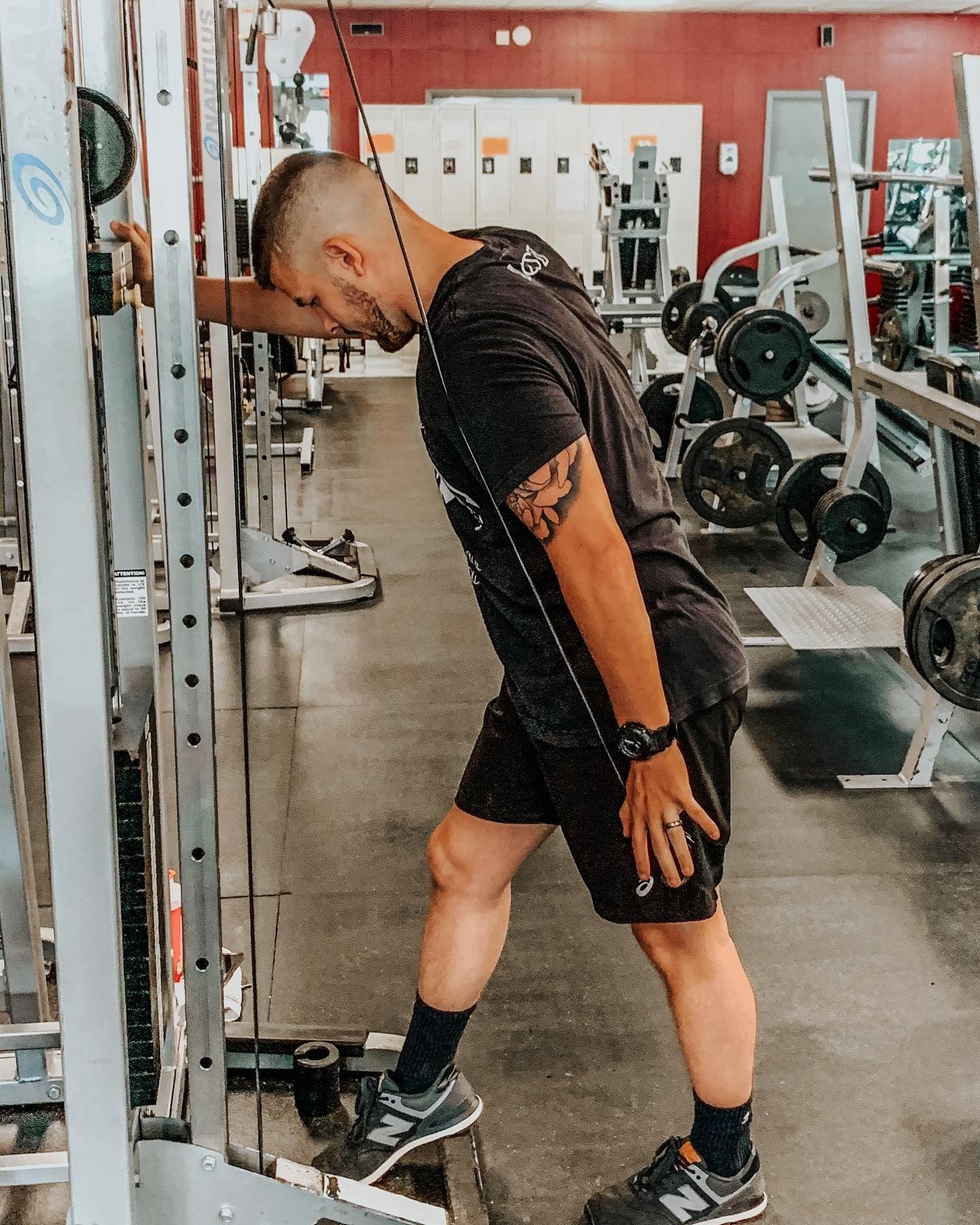 Man at a gym performing a triceps pushdown exercise, leaning toward a cable machine. He wears black workout clothes, and his right hand is on the cable's rope.