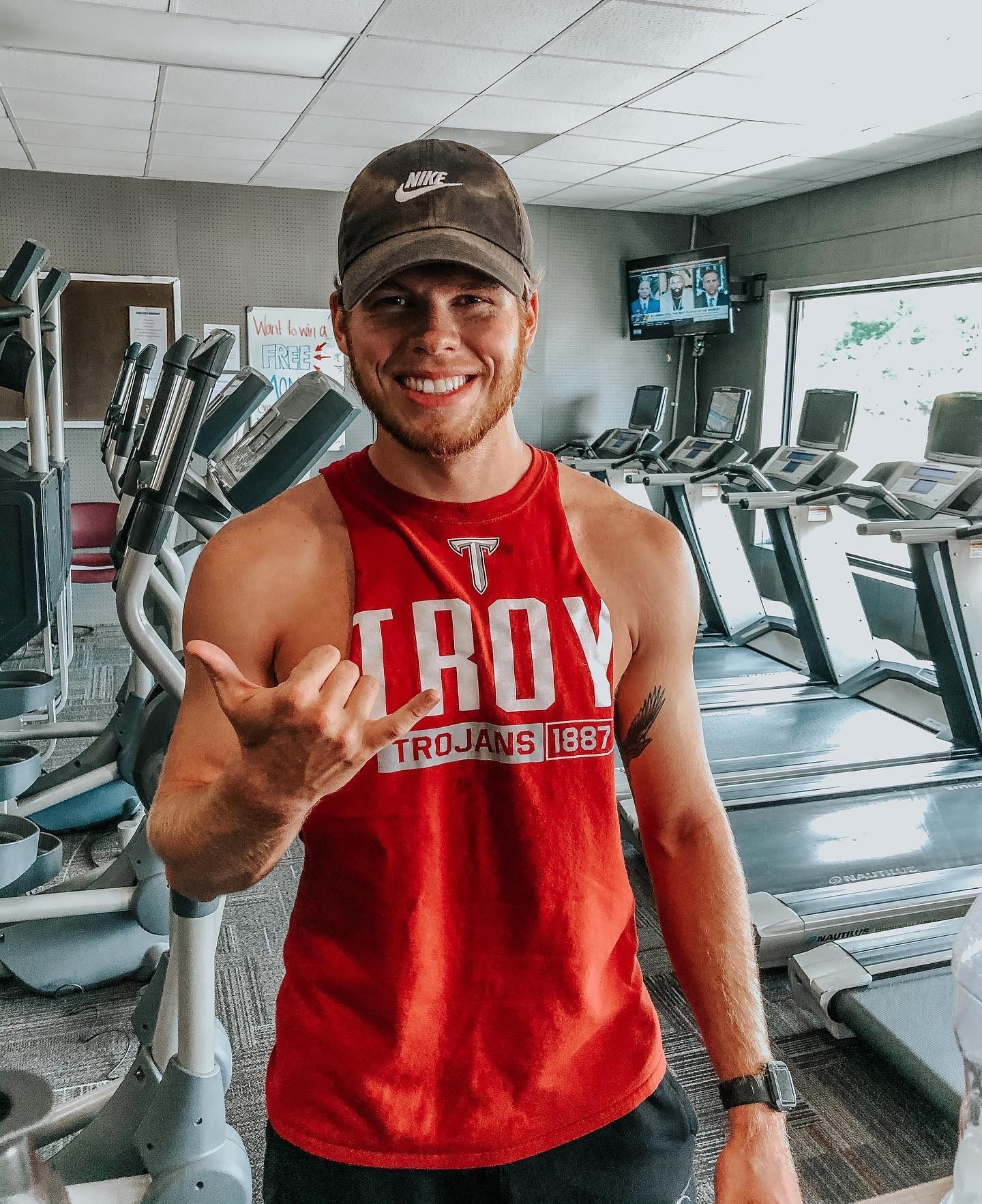Man in red tank top and baseball cap smiles, gives shaka sign in a gym with exercise equipment.